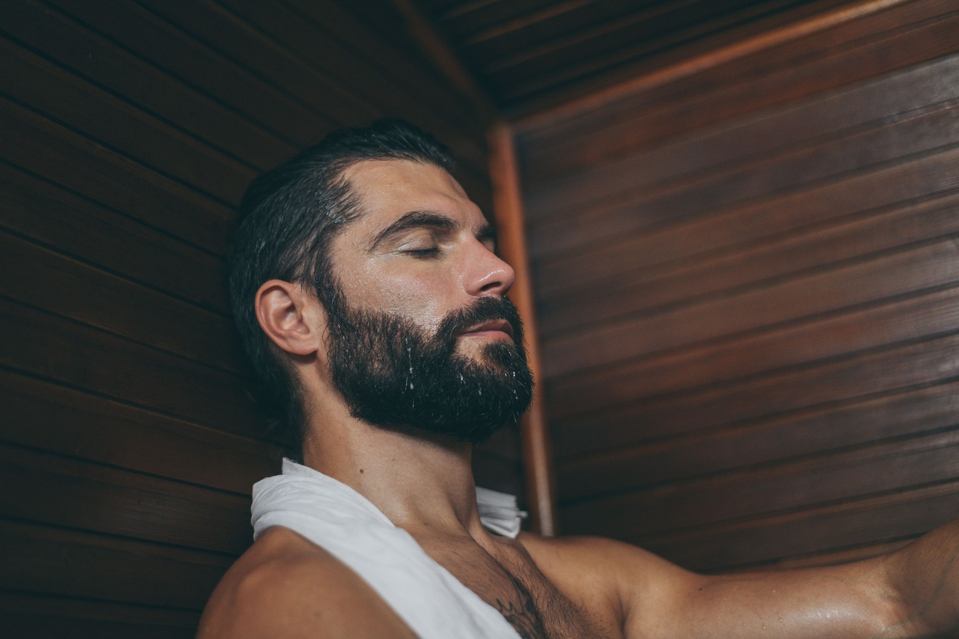 Beautiful healthy boy relaxing in a wooden sauna. Young bearded man resting after workout in the gym. Fitness male with tattoo and white towel, laying on the bench in the spa. Wellness, health concept